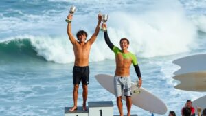Surfers Lelior and Piter celebrate winning trophies on a podium.