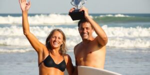 Surfers celebrate winning trophies on a sunny beach.